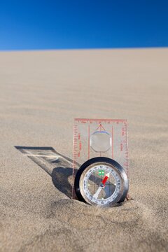 Orienteering compass placed upright in a sand dune beneath a clear blue sky, symbolizing navigation and exploration.