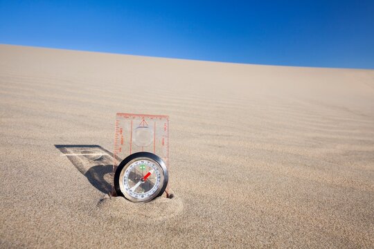Orienteering compass placed upright in a sand dune beneath a clear blue sky, symbolizing navigation and exploration.