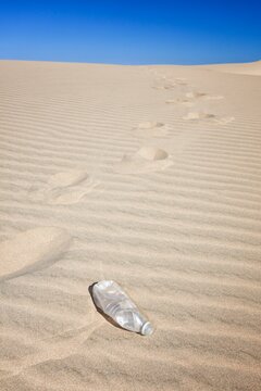 Empty plastic water bottle on rippled sand dune with footprints trailing into the distance under a clear blue sky.