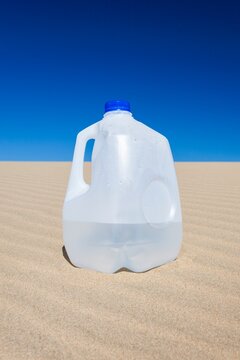 Plastic gallon water jug resting on sunlit sand dune with a vibrant blue sky overhead. Survival and hydration concept in an arid environment.