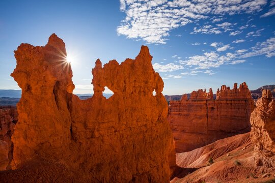 A dramatic hoodoo wall pierced with natural windows catches the sunburst at sunrise in Bryce Canyon National Park, with deep red rock and a crisp blue sky overhead.