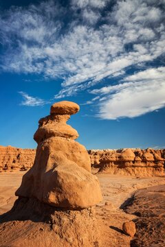 A tall hoodoo rock formation rises from the desert floor in Goblin Valley State Park, Utah, surrounded by unique sandstone shapes under a vivid blue sky with scattered clouds.