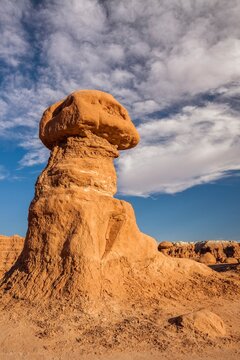 Tall hoodoo rock formation in Goblin Valley State Park, Utah, standing against a deep blue sky with drifting white clouds in the desert landscape, unique sandstone structures in warm red rocks.