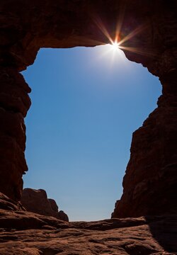 Dramatic view of the sunburst shining through a natural rock arch (Turret Arch) in Arches National Park, Utah, with deep shadows contrasting against a bright blue sky.