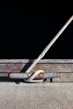 Close-up of a braided rope tied to a weathered and rusted dock cleat, set against aged wooden planks and dark water.