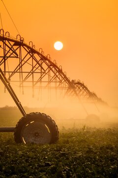 A modern center-pivot irrigation system waters a crop under a hazy orange sunset