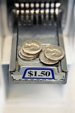 Close-up of stacks of United States coins, quarters on the sliding payment tray of a coin-operated washing machine in a laundromat, showing a $1.50 price label and chrome details.