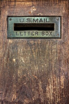Close up of an antique U.S. Mail letter slot set in a worn wooden door, showing brass patina and wood grain texture for a nostalgic postal theme.