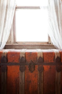 Antique steamer trunk sits beneath a bright window with lace curtains. Warm sunlight and rustic textures create a nostalgic farmhouse interior.