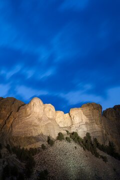 Mount Rushmore illuminated at dusk beneath a deep blue sky with soft cloud motion, carved granite faces above pines in the Black Hills of South Dakota. No people.