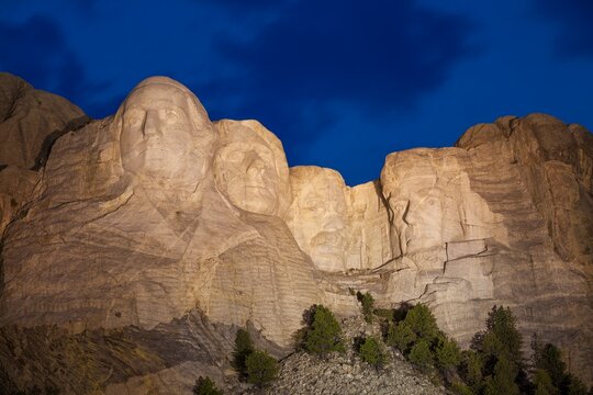 Mount Rushmore illuminated at dusk beneath a deep blue sky with soft cloud motion, carved granite faces above pines in the Black Hills of South Dakota. No people.