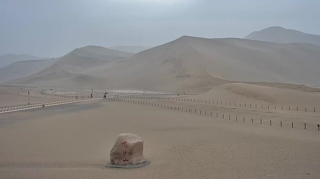 The majestic singing sand dunes of Mingsha Mountain in Dunhuang, Gansu. A serene landscape featuring smooth sand curves and a stone marker under a soft overcast sky.