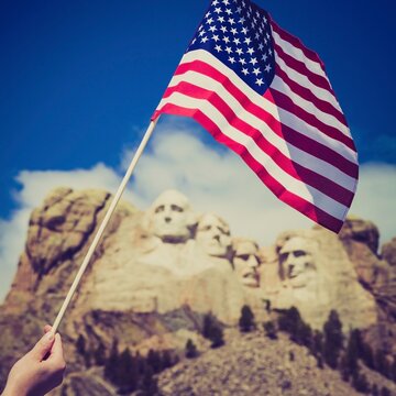 A person waves a United States flag before the granite presidents of Mount Rushmore on a bright day in the Black Hills of South Dakota.