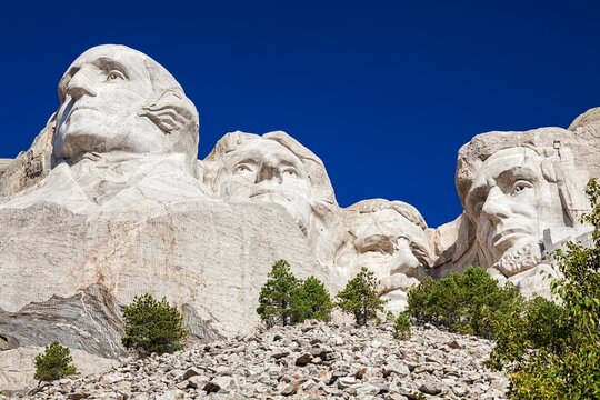 Granite carvings of Washington, Jefferson, Roosevelt, and Lincoln at Mount Rushmore National Memorial rise above pines under a clear blue sky in the Black Hills of South Dakota.