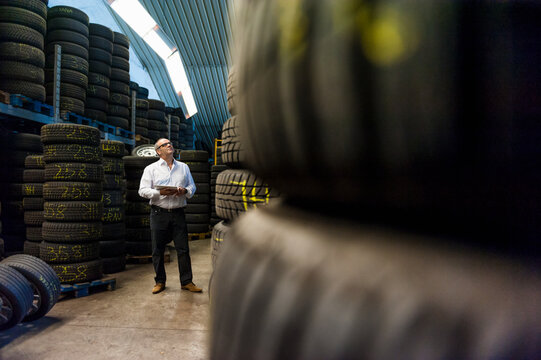 Senior businessman looking up while standing with digital tablet amidst tires