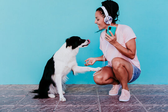 Smiling woman listening music through headphones while doing handshake with Border Collie dog against turquoise wall