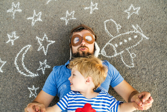 Portrait of mature man wearing pilot hat and his little son lying on asphalt painted with airplane, moon and stars