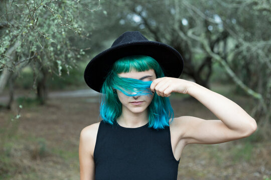 Portrait of young woman with dyed blue and green hair wearing black hat on rainy day