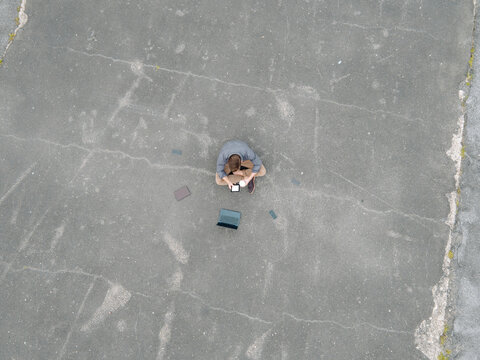 Young businessman with coffee to go and several mobile devices sitting on tarmac using tablet, quadcopter view