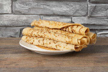 Stack of thin crepes on a white plate placed on a wooden table next to a stone wall in a kitchen setting during daylight hours