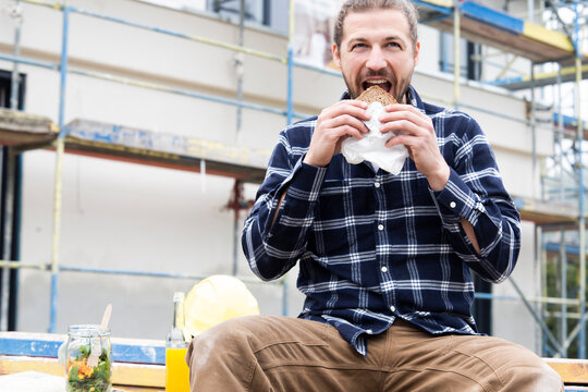 Construction worker eating bread while sitting against building at construction site