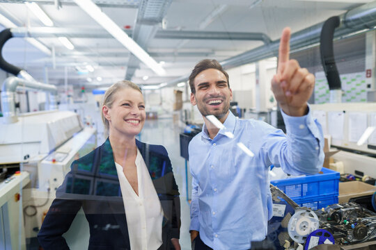 Happy young male technician pointing at graphical interface on glass in factory