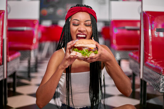 Young woman with braided hairstyle sitting on the floor and grabbing a big hamburger with her two hands