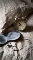 Vintage twin bell alarm clock on rumpled linen pillow with sleep mask and earplugs for quiet bedtime routine