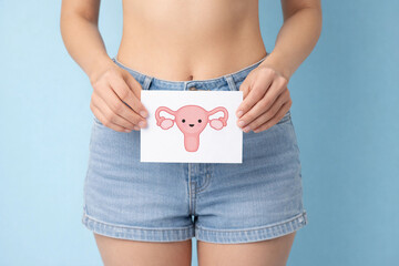 Woman holding card with cute uterus illustration in front of lower abdomen on pastel blue background, reproductive health awareness concept, medical education and women care theme.