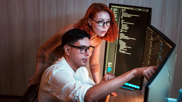 Two software developers collaborate at dual monitors, reviewing code in a dim office; one points at the screen while the other observes attentively.