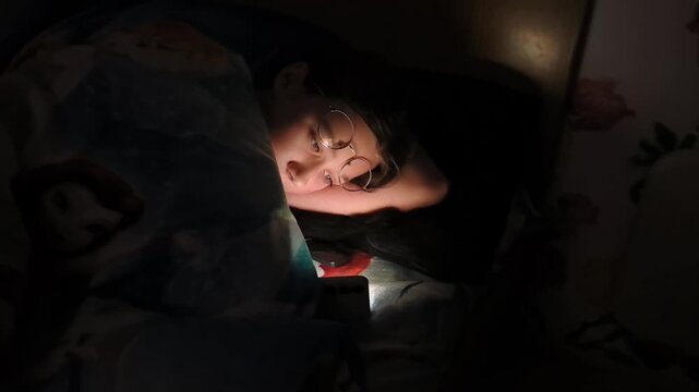 A child is in bed covered with a blanket while using a flashlight to read a book at night. The room is dark, creating a focused atmosphere for reading.