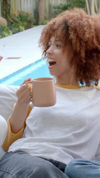 Vertical video: Reacting woman in yellow top gesturing after friend speaking on pool sofa with mugs