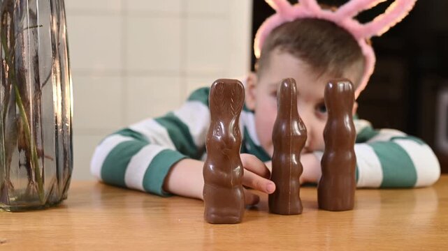 A preschool boy in a bunny costume eats a chocolate egg and a chocolate bunny at home in the kitchen, celebrating Easter. Boy playing with chocolate easter rabbit
