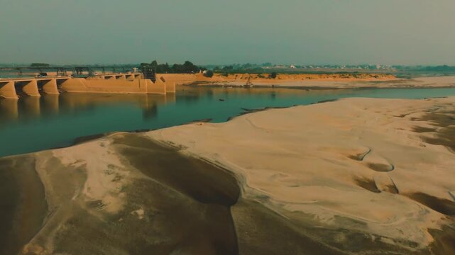 Sand bank, river water, and barrage in Sindh, Pakistan. Traffic passes over the bridge under an intense orange and teal hazy afternoon sky. Aerial left pan in 4K.