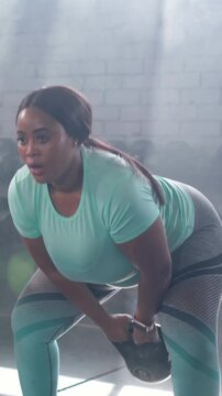 Vertical video: Holding kettlebell low mint-shirt woman swinging for fitness in gym with smartwatch