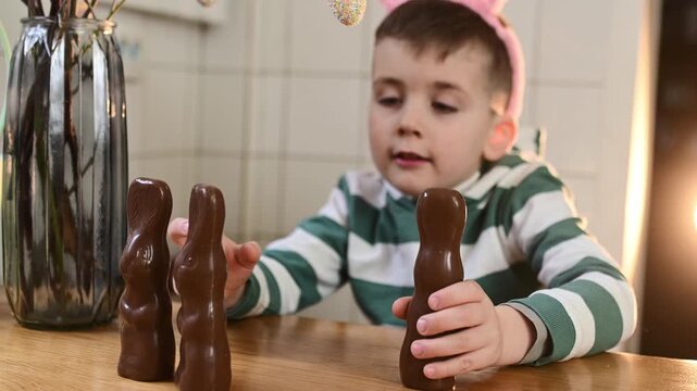 A preschool boy in a bunny costume eats a chocolate egg and a chocolate bunny at home in the kitchen, celebrating Easter. Boy playing with chocolate easter rabbit