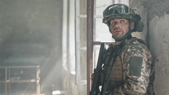 Military fighter in military uniform holding weapon during military fight. Brave man hiding from enemy attack in abandoned buildings. Armed forces concept. Looking into sight before shooting.