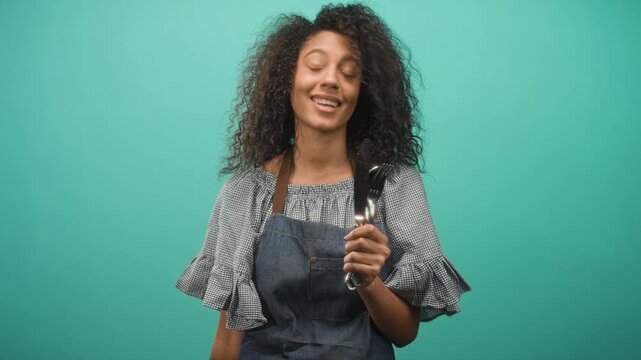 Woman holds fork and spoon in raised hand, smiling with head tilt against turquoise studio backdrop; cheerful hospitality.