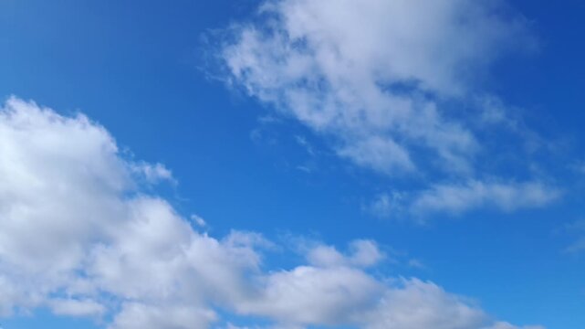 White fluffy clouds moving across a bright blue sky in real time. Beautiful real-time footage of soft white cumulus clouds drifting across a clear blue sky on a sunny day. 