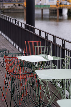 Rainy city waterfront cafe terrace with empty chairs and tables beside a railing showing outdoor dining area in a quiet documentary moment