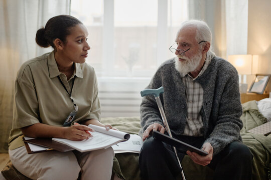 Young adult Black woman social worker holding clipboard talking with senior Caucasian man with beard sitting on bed, holding walking cane and tablet during home visit