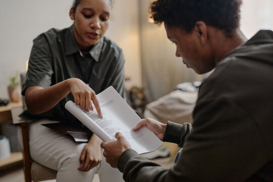 Young adult Black woman explaining documents to young zoomer during social work consultation, both seated indoors, woman gesturing toward paperwork, man attentively reading