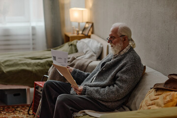 Fototapeta premium Senior Caucasian man sitting on bed checking utility bills with glasses, holding paper in both hands, appearing focused, gray hair and beard visible, bedroom setting with soft lighting
