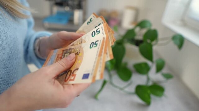 Hands counting euro banknotes on countertop, soft natural light, green pothos plant, blue knit sweater, meticulous thumb through notes, planning savings, personal finance mood Hands counting euro bank