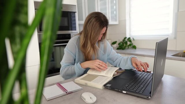 Caucasian woman typing on laptop cozy bright kitchen workspace with notebook and mouse potted plant in foreground morning light focused freelancer checking notes and sipping coffee Caucasian woman typ