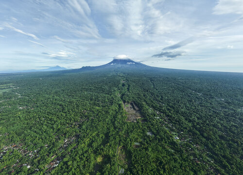 Aerial view of lush green forest stretching towards a towering mountain peak under a vast, partly cloudy sky, Klaten Regency, Jawa Tengah, Indonesia.