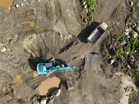 Aerial view of an excavator digging into the earth, juxtaposed with a parked truck amidst the rugged terrain of Kali woro, Klaten Regency, Jawa Tengah, Indonesia.