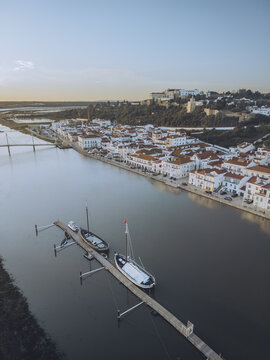 Aerial view of the tranquil Sado River reflecting the soft dawn light, with boats docked near the historic Alcacer do Sal, Setubal, Portugal.