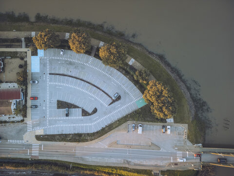 Aerial view of a geometrically designed parking lot meeting the river's edge, contrasting with the natural elements of trees and water, Alcacer do Sal, Setubal, Portugal.
