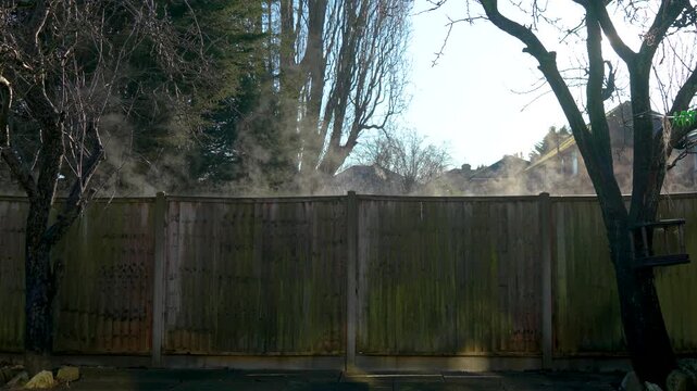 Fire emergency unfolding in private sector residential neighborhood. Thick smoke rising and drifting above wooden fence near suburban houses. Accident scene escalating while sunlight piercing through